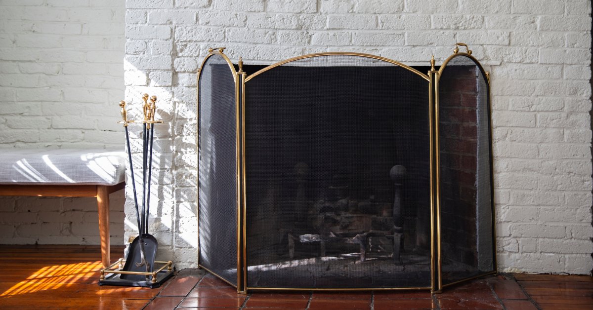An unlit, white brick fireplace with a golden and black mesh screen with a golden-handled fire poker and shovel next to it.