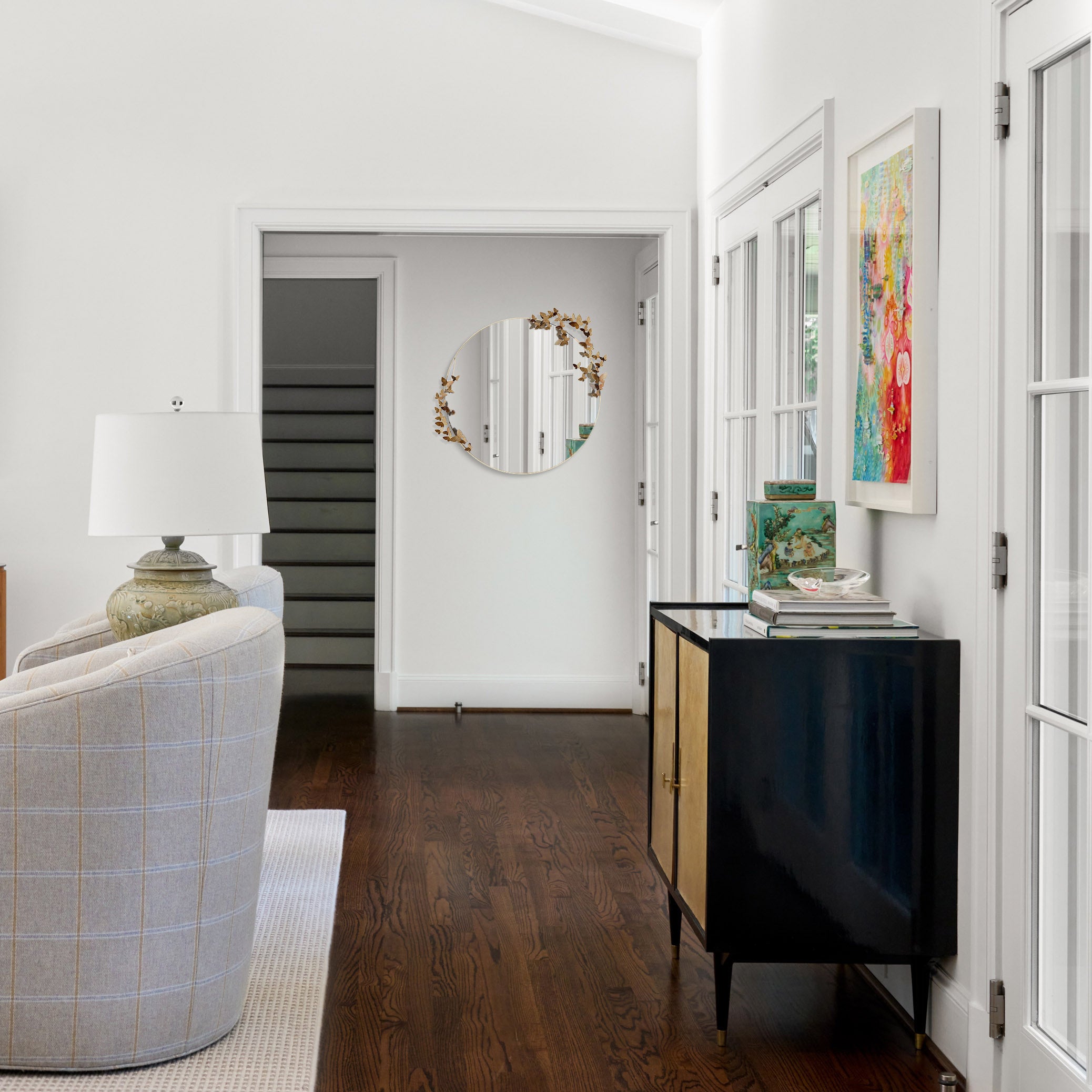 A hallway with a luxury round butterfly mirror on a white wall, a black and gold console table holding colorful decor, and a cozy armchair by a lamp.