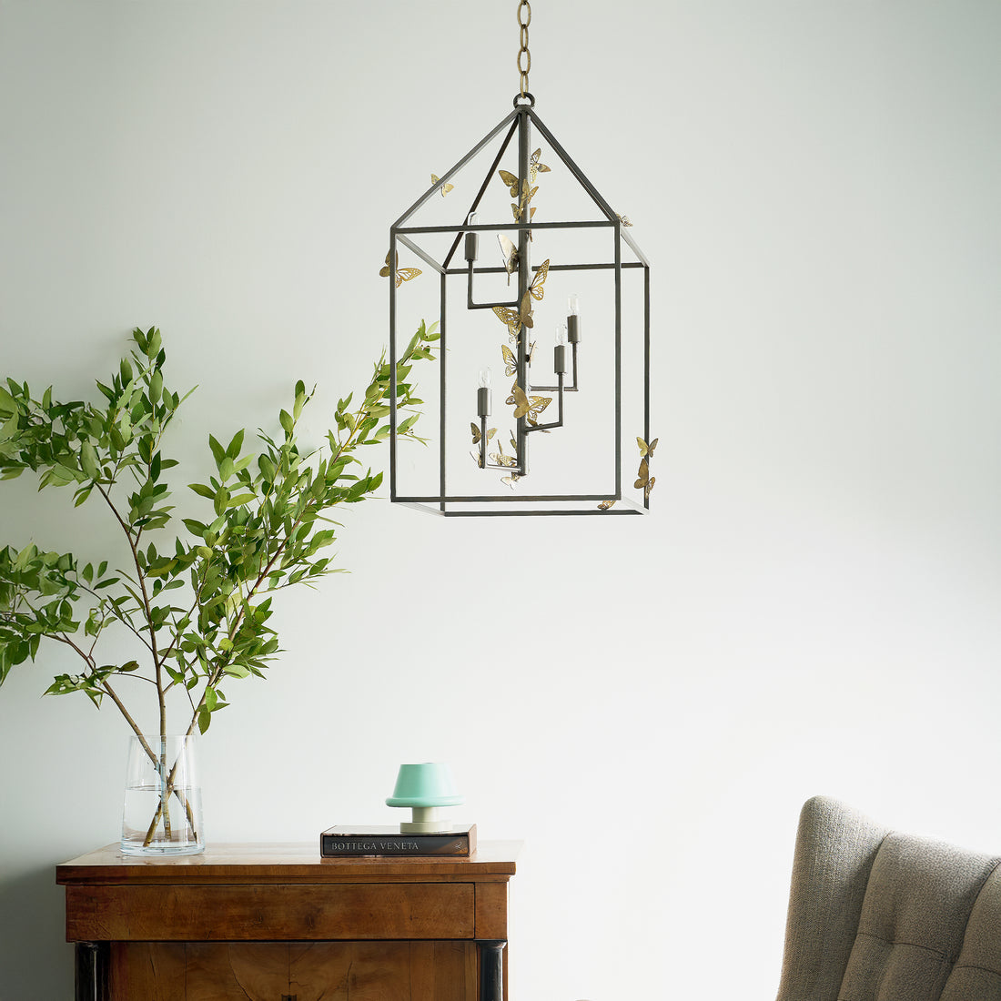 Monarch lantern chandelier with a house shaped frame featuring metal butterfly accents, housing multiple lightbulbs. Hanging by a chain.