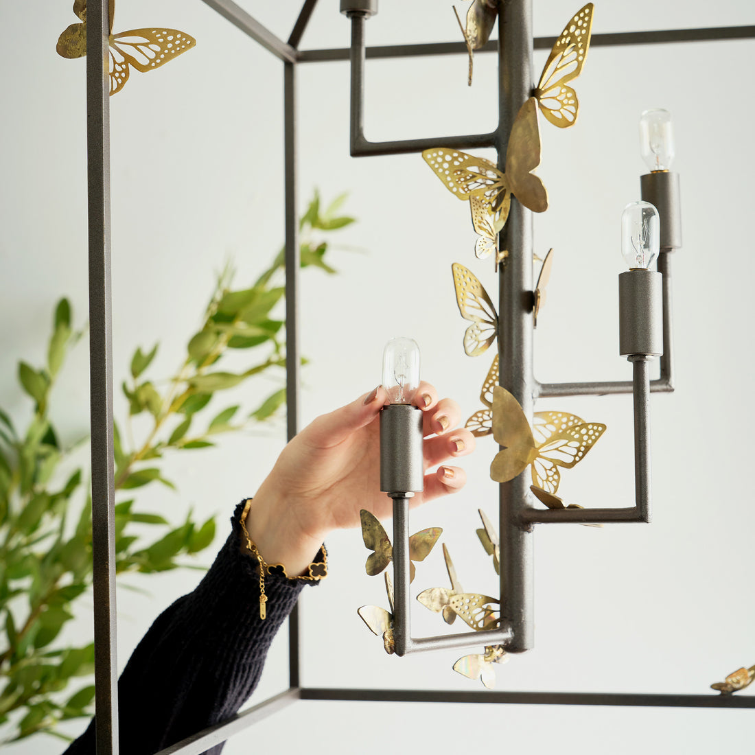 Hand adjusts light bulb on a monarch lantern adorned with metal butterflies. Metallic fixture contrasts with delicate gold accents.