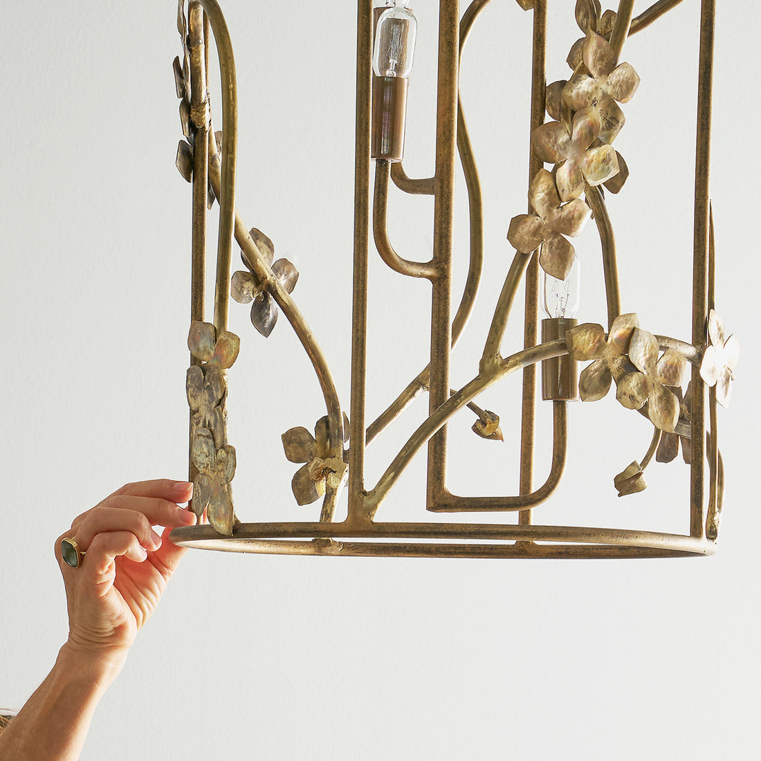 A hand adjusts a decorative sakura lantern chandelier with floral designs in a brass finish, highlighting its intricate, organic metalwork.
