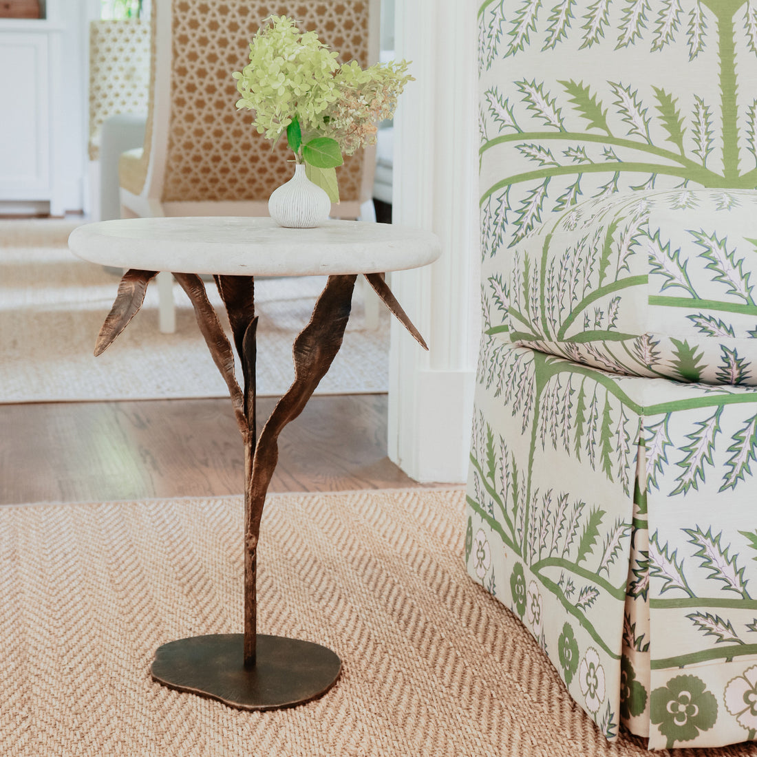 Elegant willow accent table by Claire Crowe Collection with a marble top and metal base, next to a patterned green and cream upholstered chair.