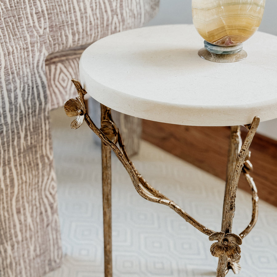 Luxury cherry blossom accent table with a white stone top and bronze branch design legs, placed on a geometric rug next to a textured chair.