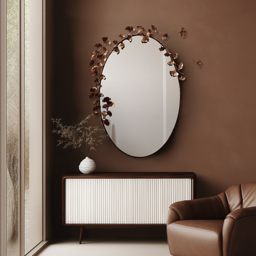 Custom ginkgo mirror with metal leaf accents on a brown wall, above a ribbed front sideboard and next to a leather chair, creating a cozy atmosphere.